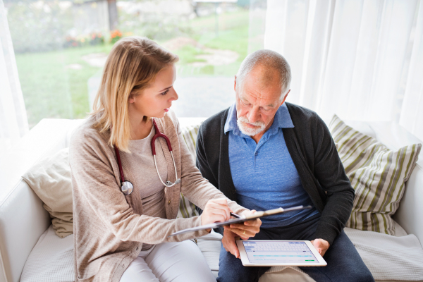 Health visitor and a senior man during home visit. A female nurse or a doctor showing test results on a tablet.