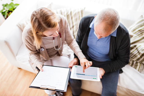 Health visitor and a senior man during home visit. A female nurse or a doctor showing test results on a tablet. High angle view.