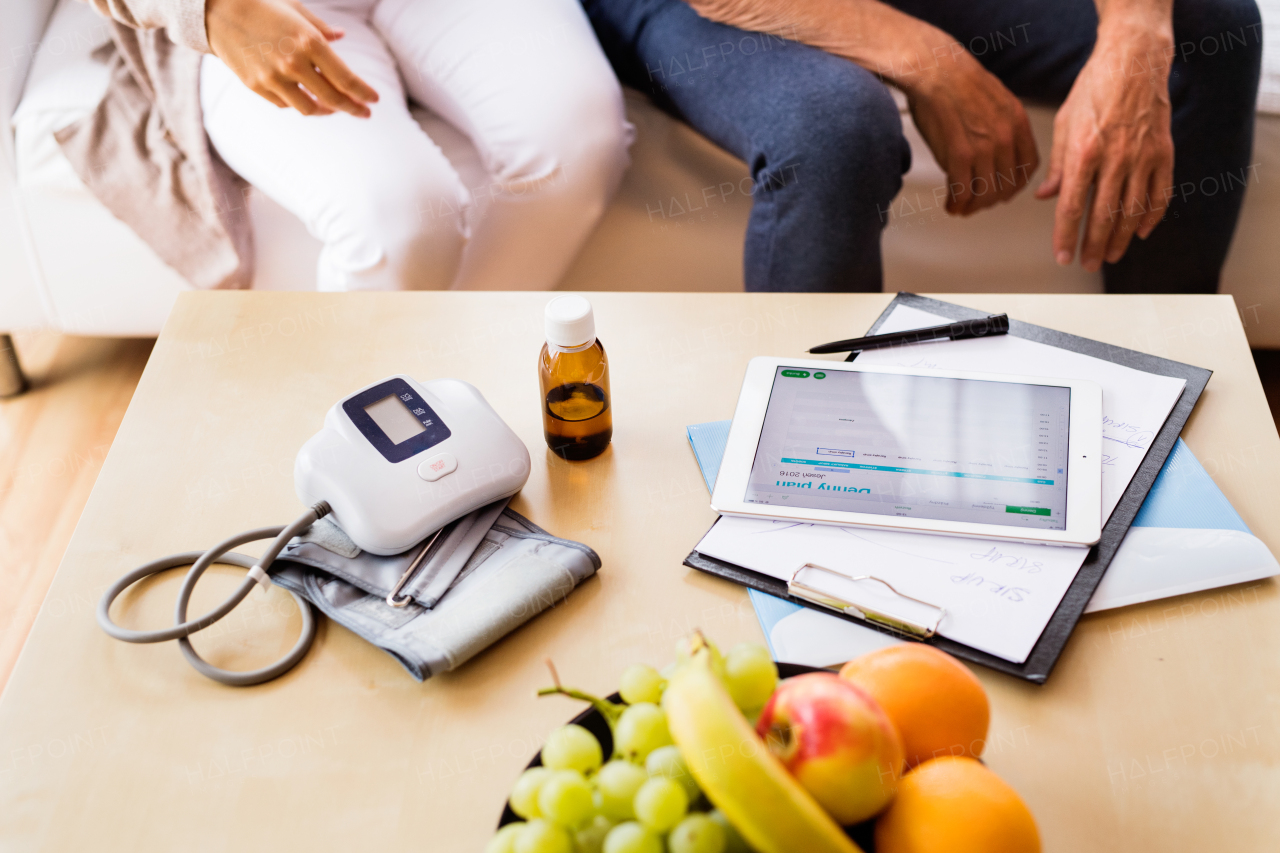 Unrecognizable health visitor and a senior man during home visit. A female nurse or a doctor showing test results on a tablet.