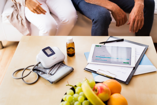 Unrecognizable health visitor and a senior man during home visit. A female nurse or a doctor showing test results on a tablet.