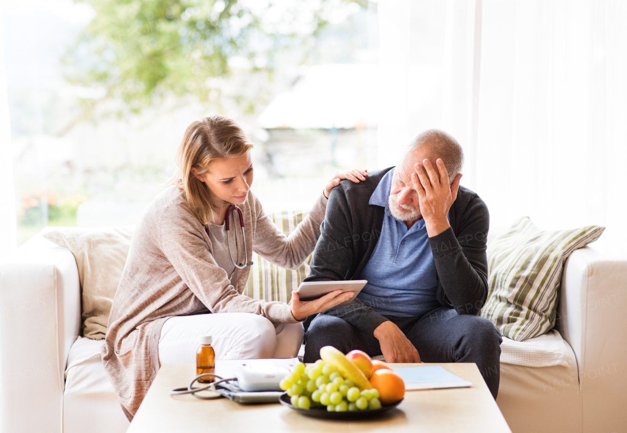 Health visitor and a senior man during home visit. A female nurse or a doctor showing test results on a tablet. Man feeling disappointed, covering face with hands.