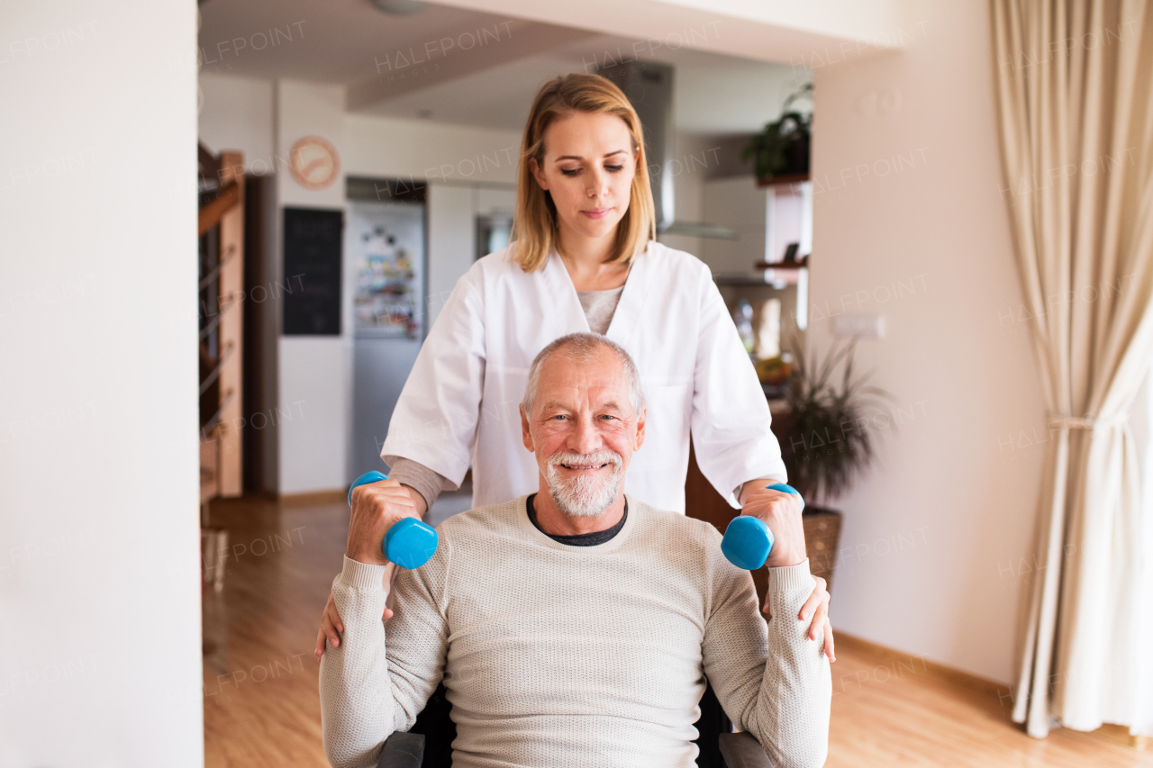 Health visitor and a senior man in a wheelchair during home visit. A nurse or a physiotherapist helping a senior man exercise with dumbbells.