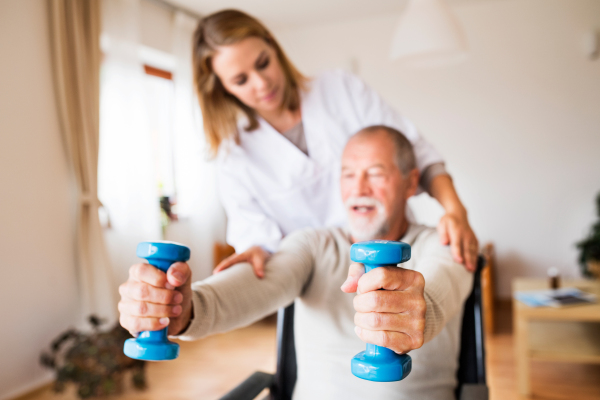 Health visitor and a senior man in a wheelchair during home visit. A nurse or a physiotherapist helping a senior man exercise with dumbbells.