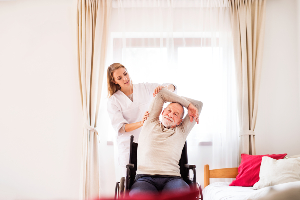 Health visitor and a senior man in a wheelchair during home visit. A nurse or a physiotherapist helping a senior man exercise.