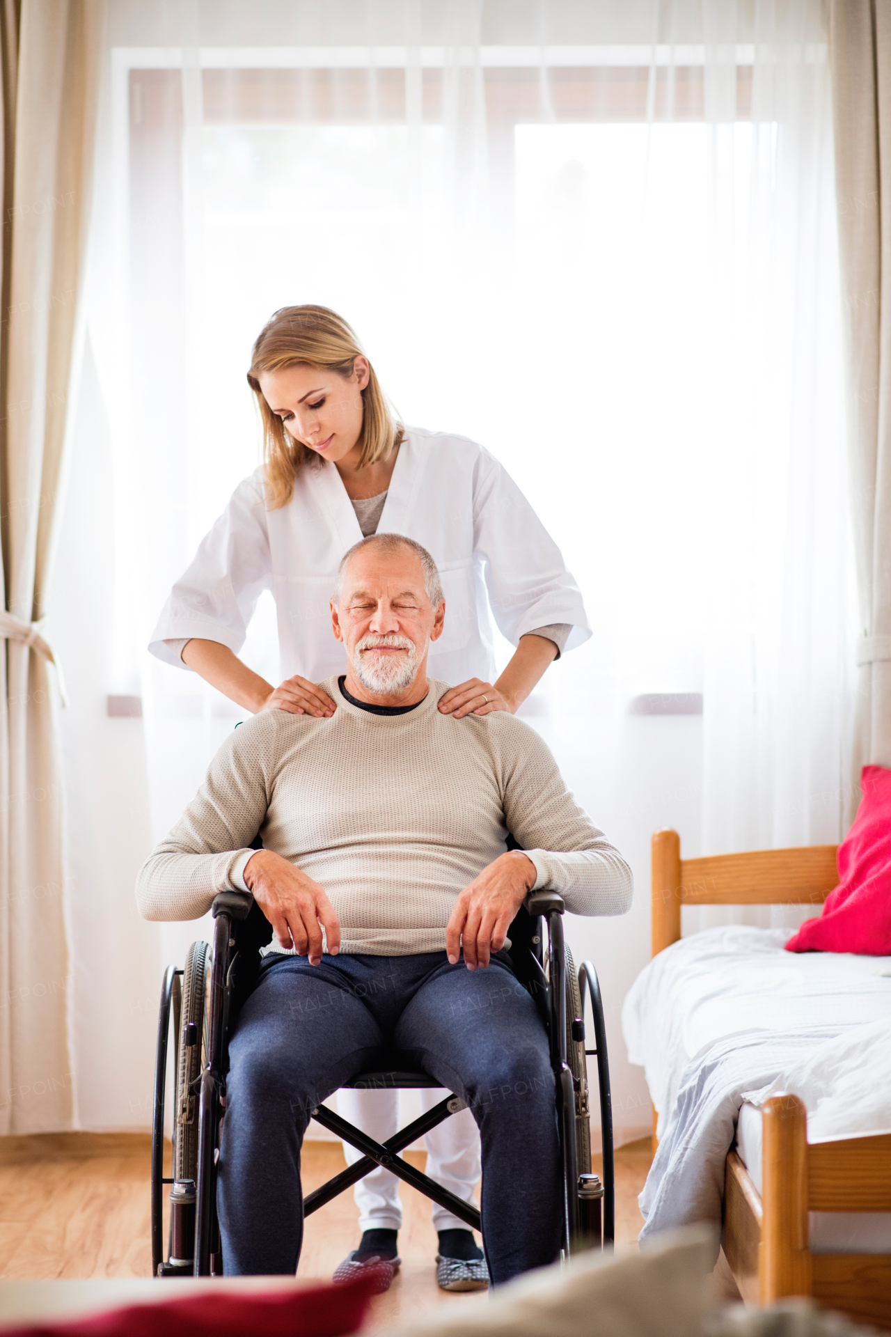 Health visitor and a senior man in a wheelchair during home visit. A nurse or a physiotherapist giving man shoulder massage.