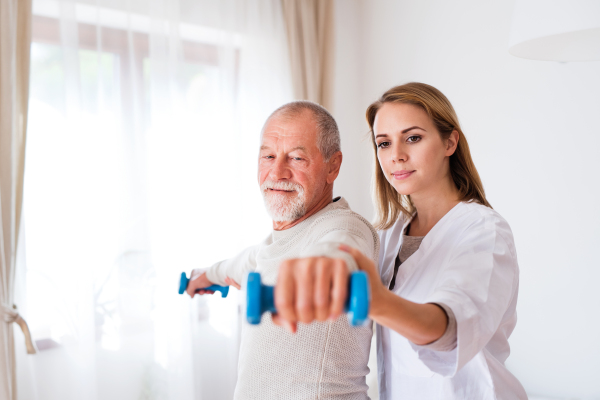 Health visitor and a senior man during home visit. A nurse or a physiotherapist helping a senior man exercise with dumbbells.