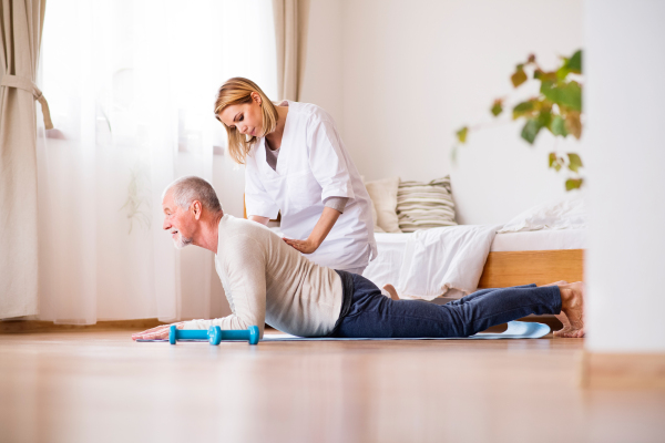 Health visitor and a senior man during home visit. A nurse or a physiotherapist helping a senior man exercise.