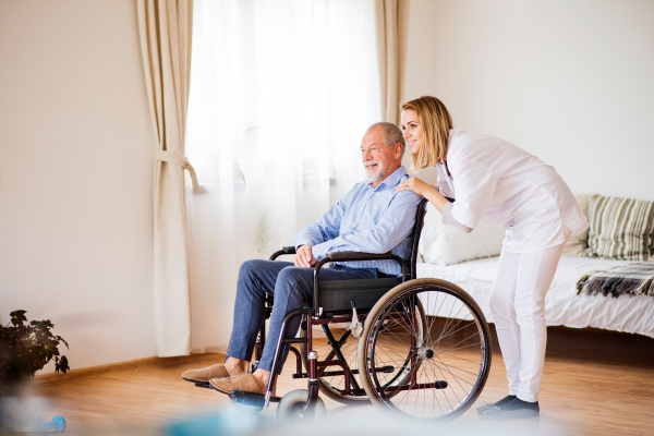 Health visitor or a nurse and a senior man in a wheelchair during home visit. Man and woman looking out of a window.