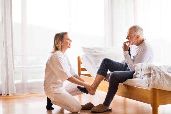 Health visitor and a senior man during home visit. A nurse helping a senior man to put socks on.