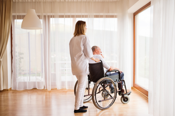 Health visitor or a nurse and a senior man in a wheelchair during home visit. Man and woman looking out of a window.