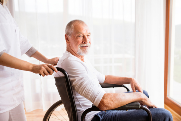 Unrecognizable health visitor or a nurse and a senior man in a wheelchair during home visit.