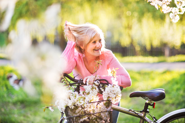 Beautiful senior woman with bicycle standing outside in spring nature under blossoming trees.