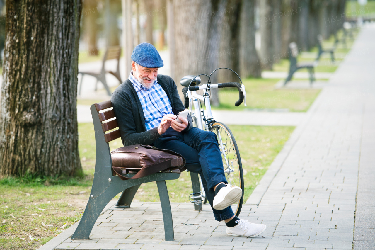 Handsome senior man with bicycle in town park sitting on bench, holding smart phone, texting. Sunny spring day.