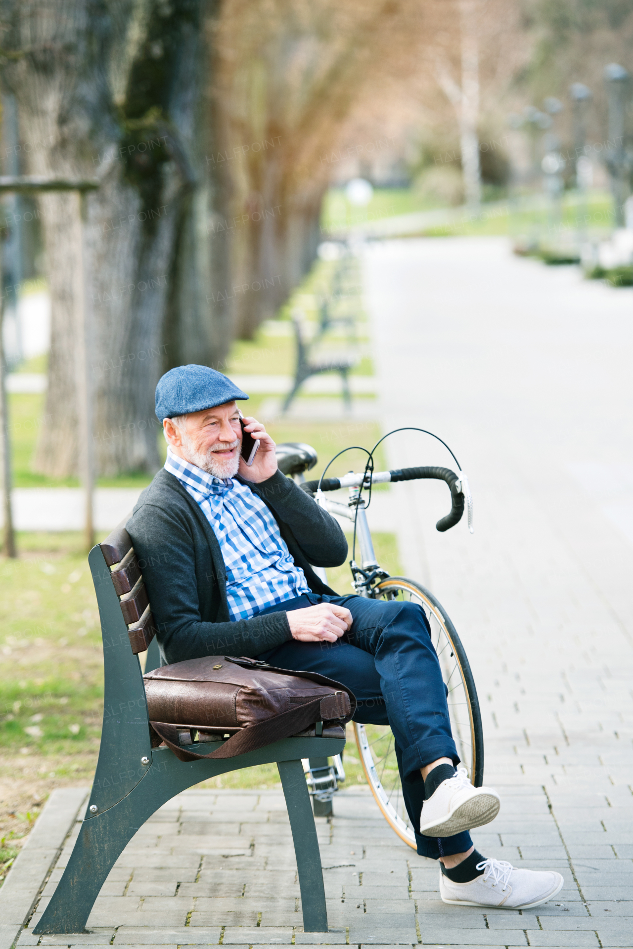 Handsome senior man with bicycle in town park sitting on bench with legs crossed, holding smart phone, making phone call. Sunny spring day.