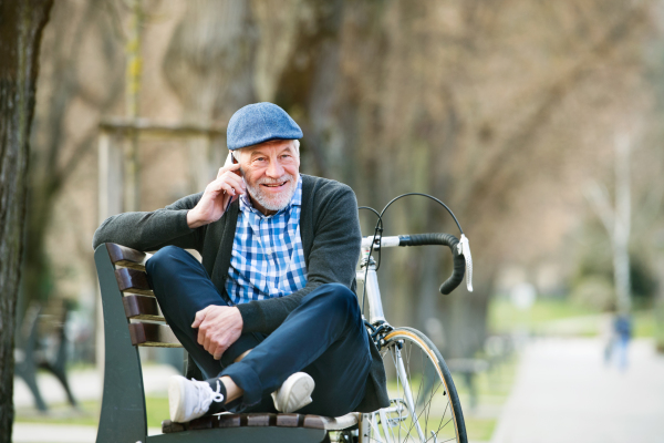 Handsome senior man with bicycle in town park sitting on bench with legs crossed, holding smart phone, making phone call. Sunny spring day.