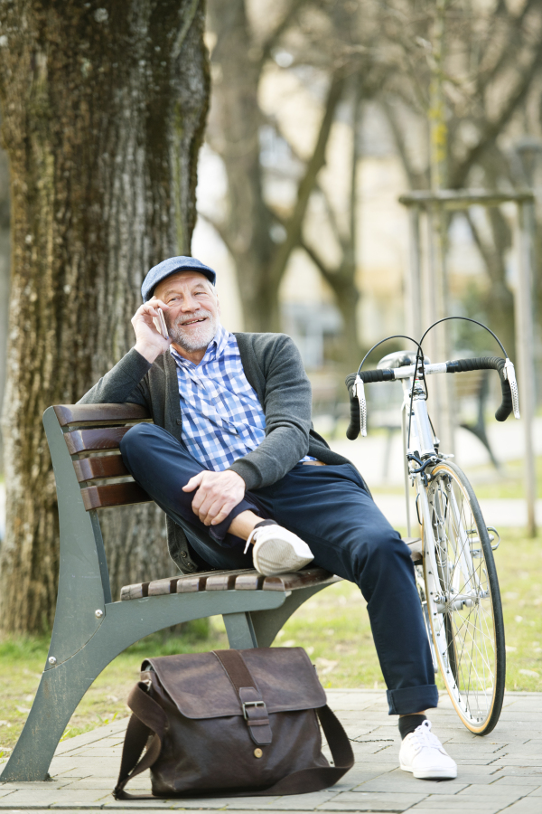 Handsome senior man with bicycle in town park sitting on bench, holding smart phone, making phone call. Sunny spring day.