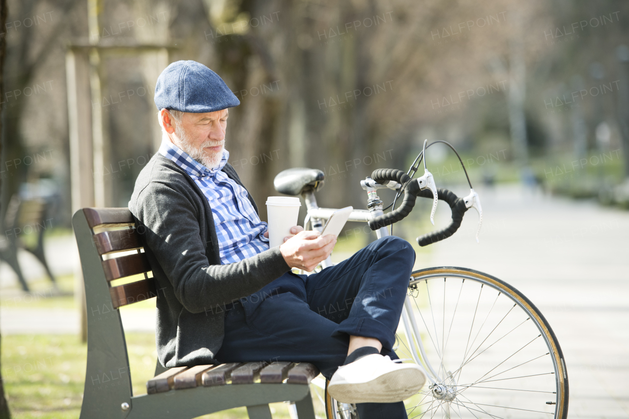 Handsome senior man with bicycle in town park sitting on bench, holding coffee and smart phone, texting. Sunny spring day.