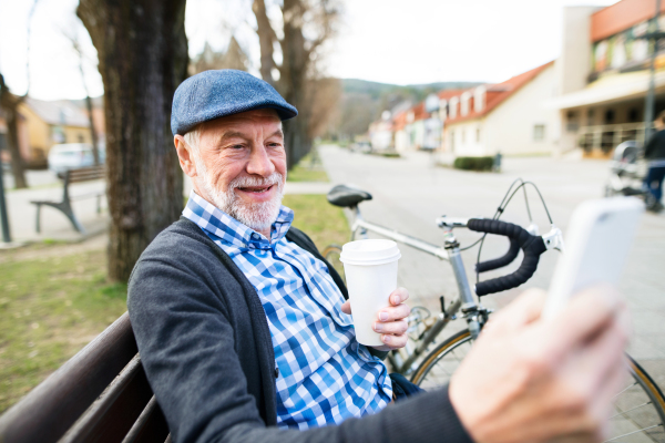 Handsome senior man with bicycle in town park sitting on bench, drinking coffee, holding smart phone, taking selfie. Sunny spring day.