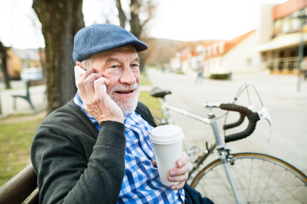 Handsome senior man with bicycle in town park sitting on bench, holding coffee and smart phone, making phone call. Sunny spring day.