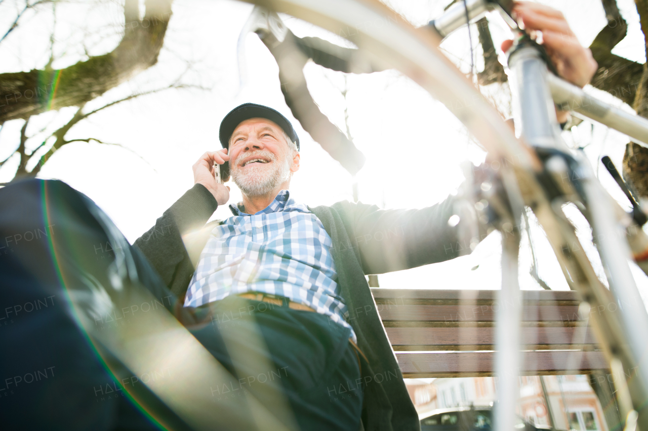 Handsome senior man with bicycle in town park sitting on bench with legs crossed, holding smart phone, making phone call. Sunny spring day.