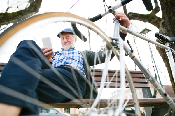 Handsome senior man with bicycle in town park sitting on bench, holding smart phone, texting. Sunny spring day.