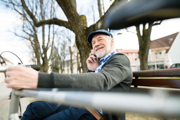 Handsome senior man with bicycle in town park sitting on bench, holding smart phone, making phone call. Sunny spring day.