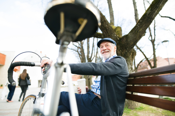 Handsome senior man with bicycle in town park sitting on bench, drinking coffee. Sunny spring day.