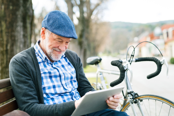 Handsome senior man with bicycle in town park sitting on bench, working on tablet. Sunny spring day.