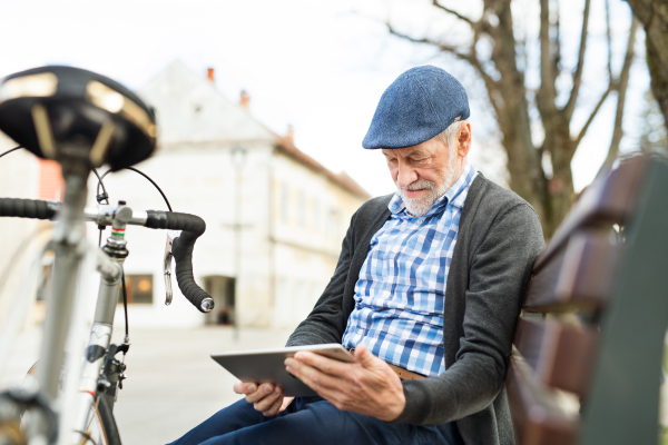 Handsome senior man with bicycle in town park sitting on bench, working on tablet. Sunny spring day.