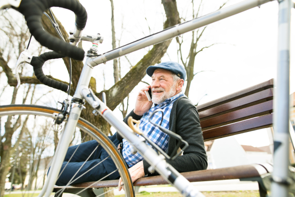 Handsome senior man with bicycle in town park lying on bench, holding smart phone, making phone call. Sunny spring day.