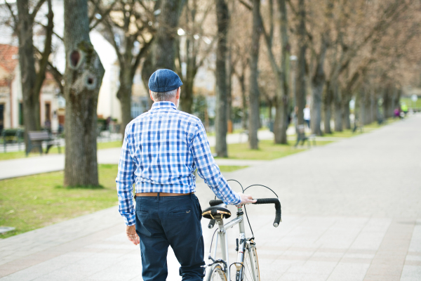 Handsome senior man in blue checked shirt with bicycle in town. Sunny spring day. Rear view.