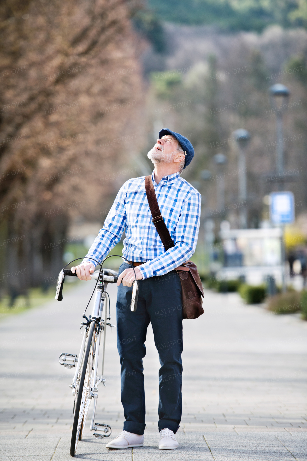 Handsome senior man in blue checked shirt with bicycle in town, looking up. Sunny spring day.