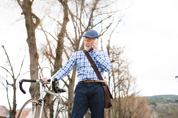 Handsome senior man in blue checked shirt with bicycle in town holding smart phone, making phone call. Sunny spring day.