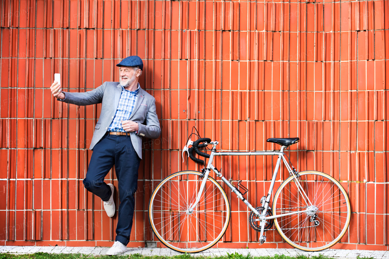 Handsome senior man in blue checked shirt and gray jacket with bicycle holding smart phone, taking selfie. Orange brick wall background.