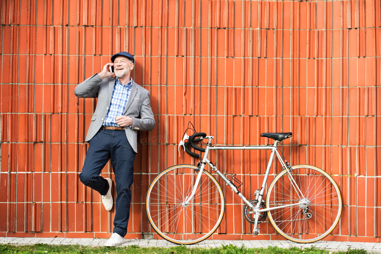 Handsome senior man in blue checked shirt and gray jacket with bicycle holding smart phone, making phone call. Orange brick wall background.