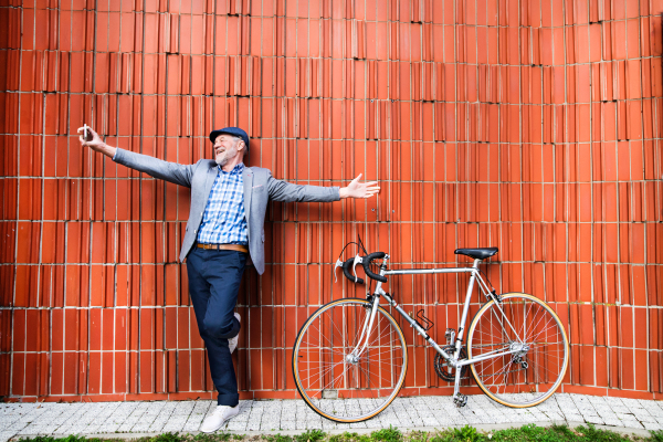 Handsome senior man in blue checked shirt and gray jacket with bicycle holding smart phone, taking selfie. Orange brick wall background.