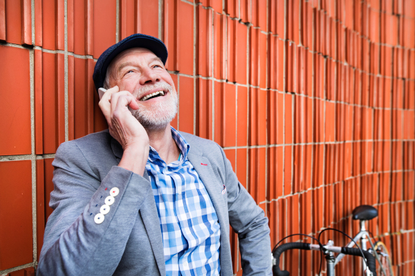 Handsome senior man in blue checked shirt and gray jacket with bicycle holding smart phone, making phone call. Orange brick wall background.