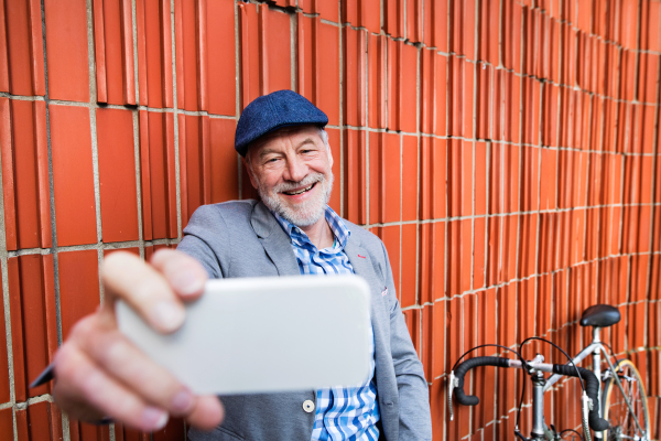 Handsome senior man in blue checked shirt and gray jacket with bicycle holding smart phone, taking selfie. Orange brick wall background.