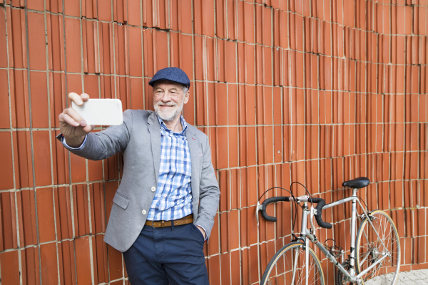 Handsome senior man in blue checked shirt and gray jacket with bicycle holding smart phone, taking selfie. Orange brick wall background.