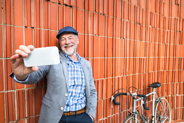 Handsome senior man in blue checked shirt and gray jacket with bicycle holding smart phone, taking selfie. Orange brick wall background.