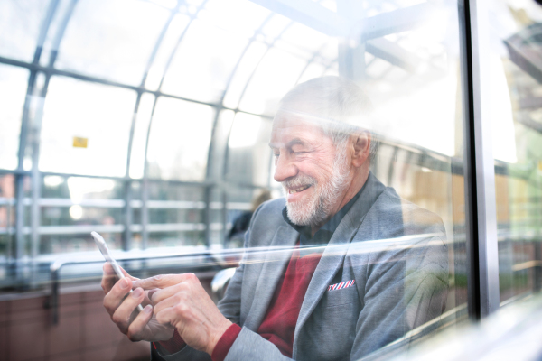 Handsome senior man in gray jacket holding smart phone, texting. Glass ceiling background. View through glass.