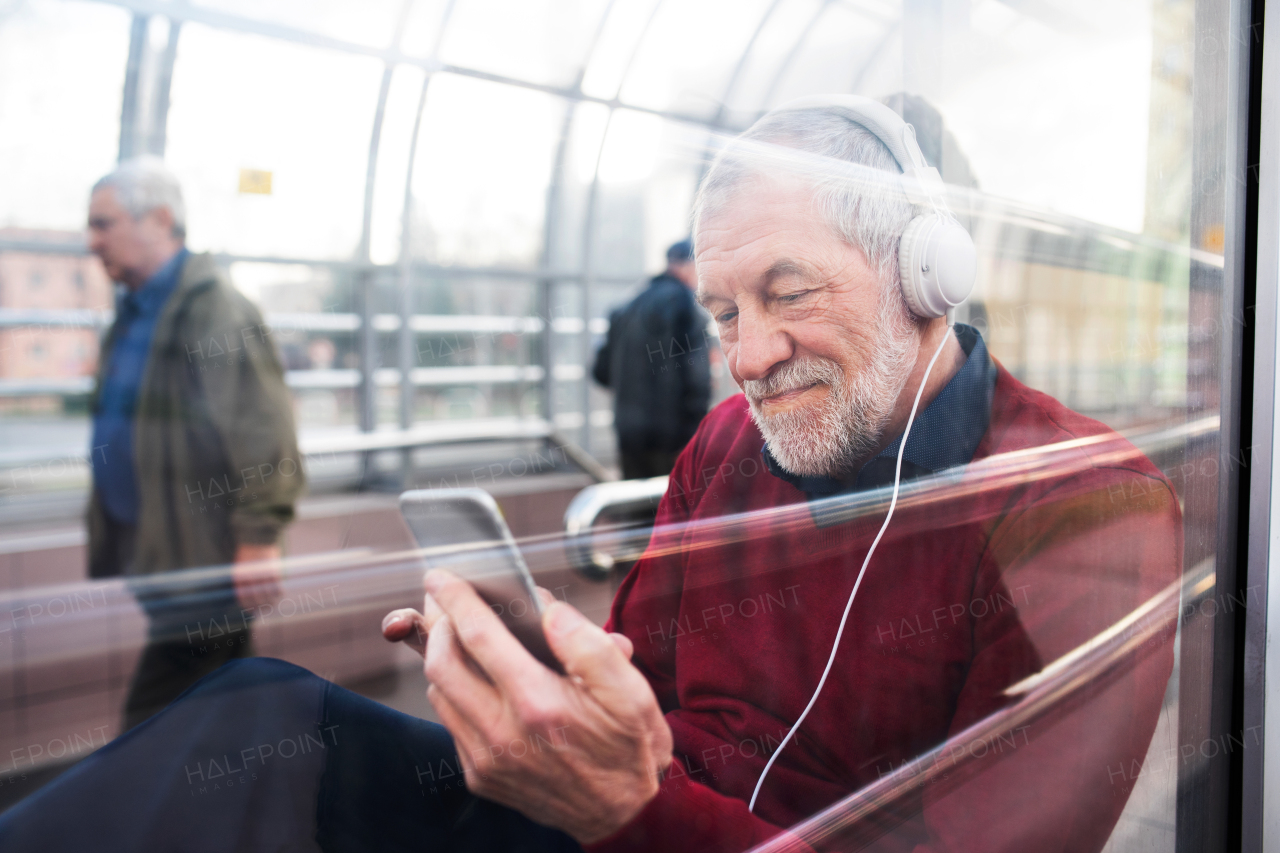 Handsome senior man holding smart phone, headphones on his ears, listening music, sitting in glass passage. View through glass.