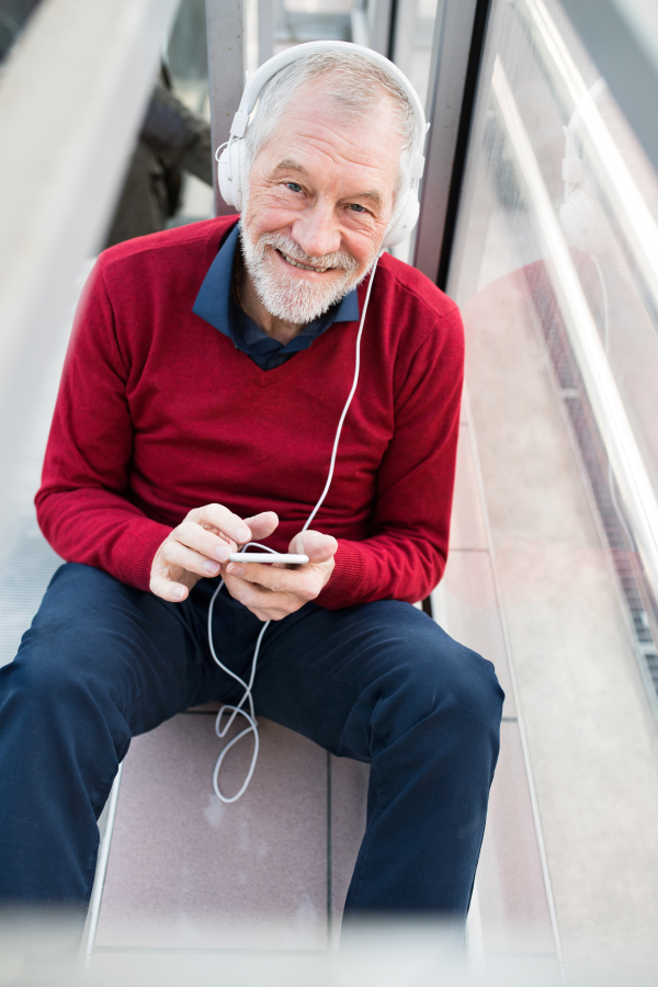 Handsome senior man holding smart phone, headphones on his ears, listening music, sitting in glass passage.