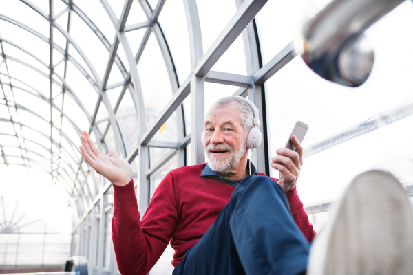 Handsome senior man holding smart phone, headphones on his ears, listening music, sitting in glass passage.