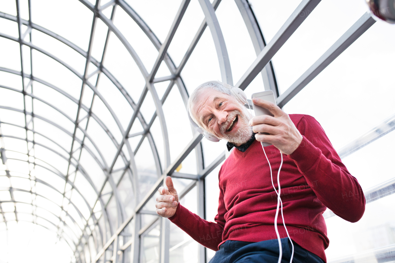 Handsome senior man holding smart phone, headphones on his ears, listening music, in glass passage.