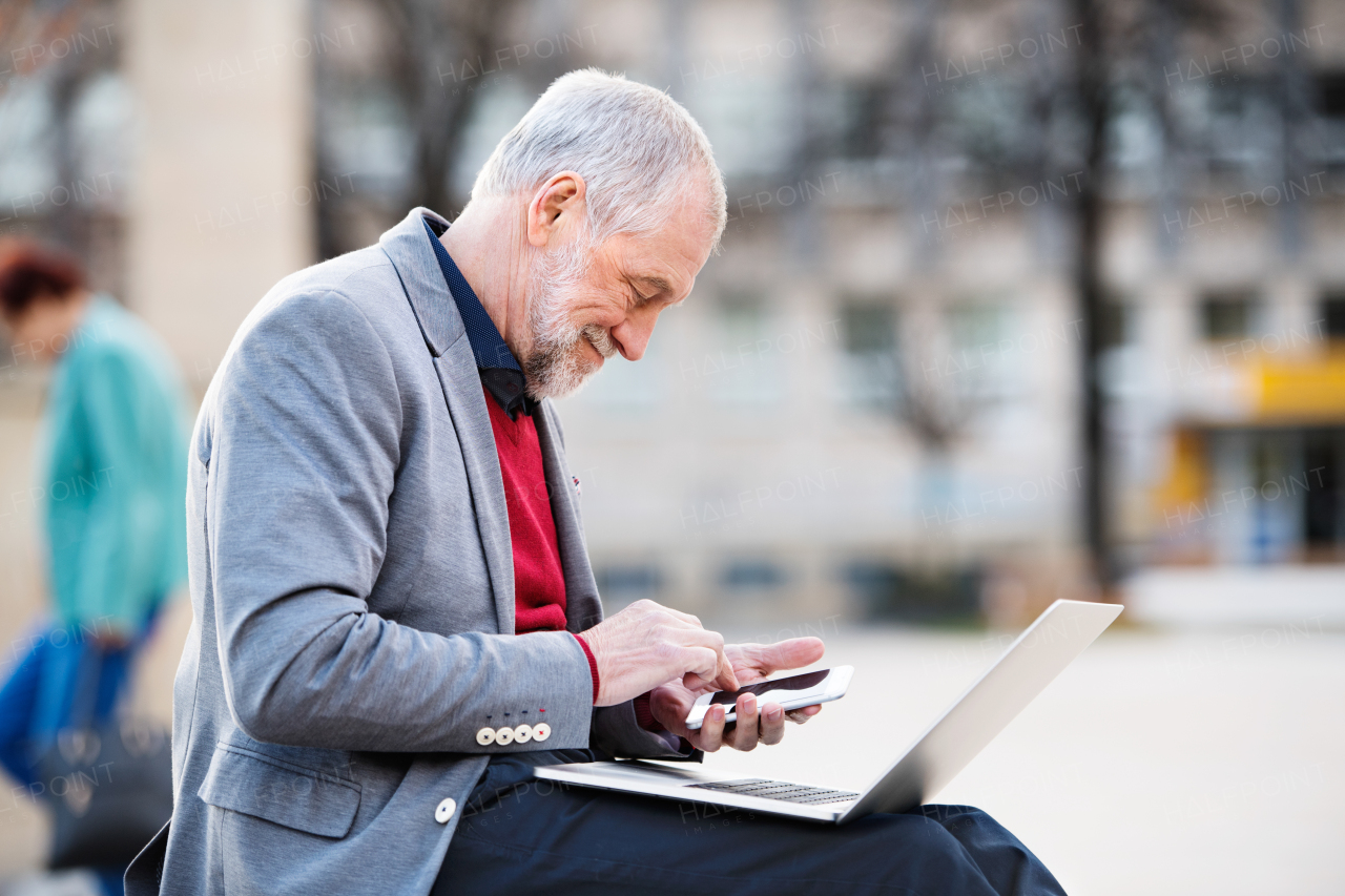 Handsome senior man in town sitting on bench, holding laptop and smart phone, making phone call. Sunny spring day.