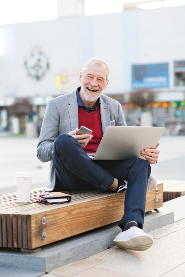 Handsome senior man in town sitting on bench, holding laptop and smart phone, making phone call. Sunny spring day.