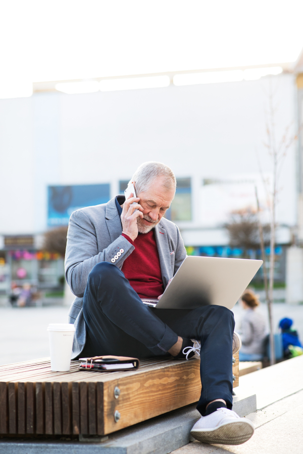 Handsome senior man in town sitting on bench, holding laptop and smart phone, making phone call. Sunny spring day.