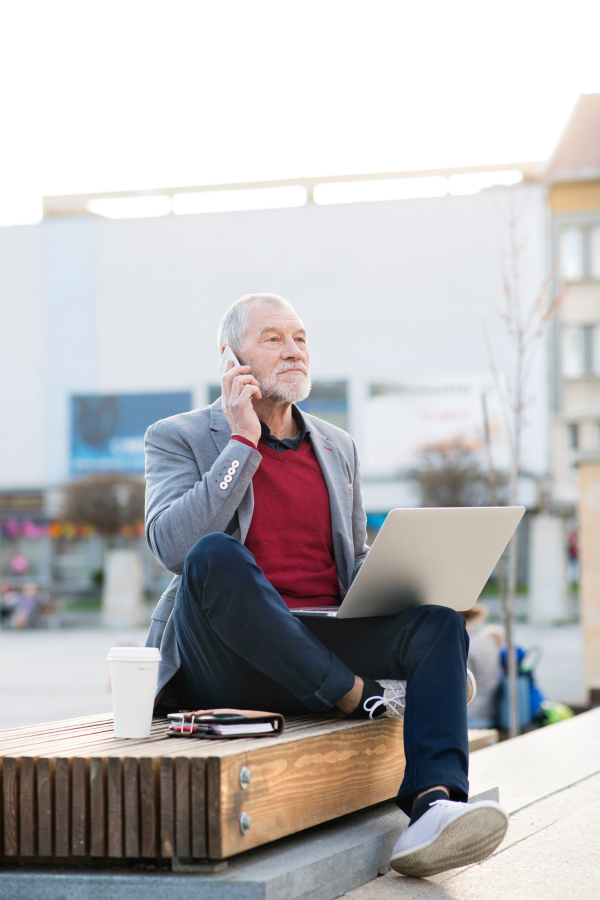 Handsome senior man in town sitting on bench, holding laptop and smart phone, making phone call. Sunny spring day.