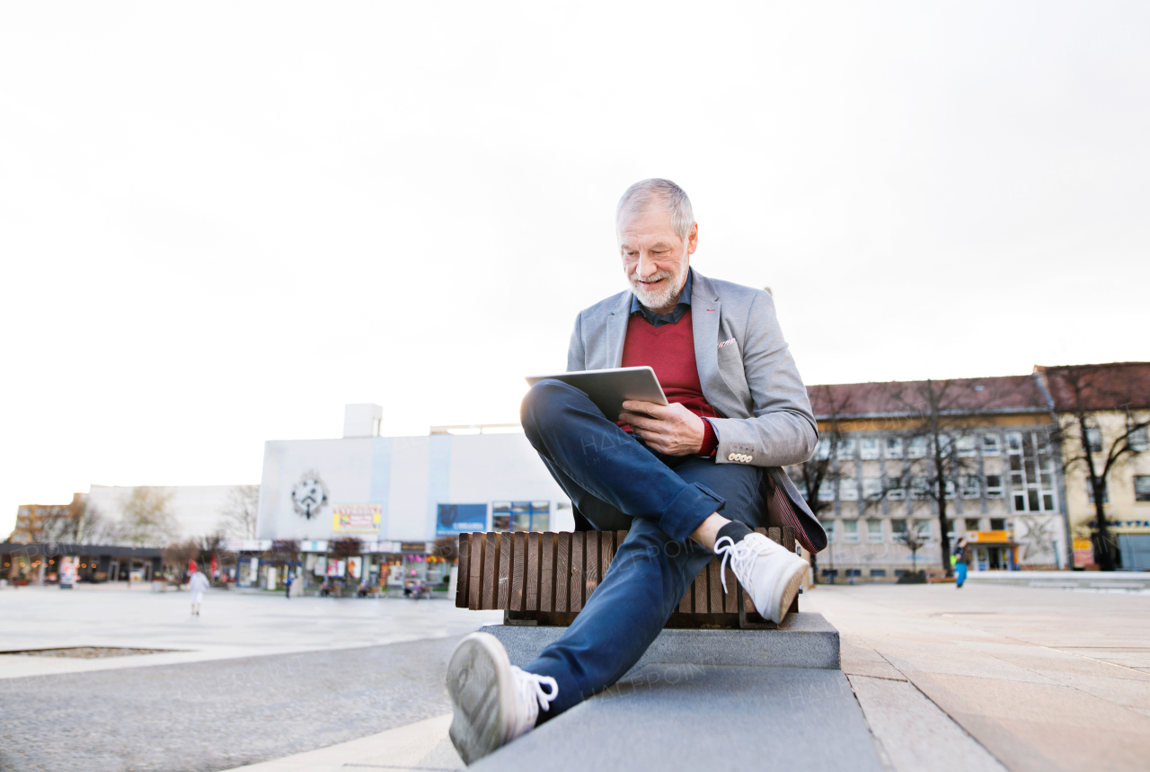 Handsome senior man in town sitting on bench, working on tablet. Sunny spring day.